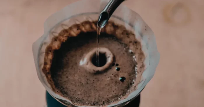 A close-up of hot water being poured from a kettle into a coffee filter filled with grounds, brewing fresh coffee in a drip coffee maker. | Hustle Coffee