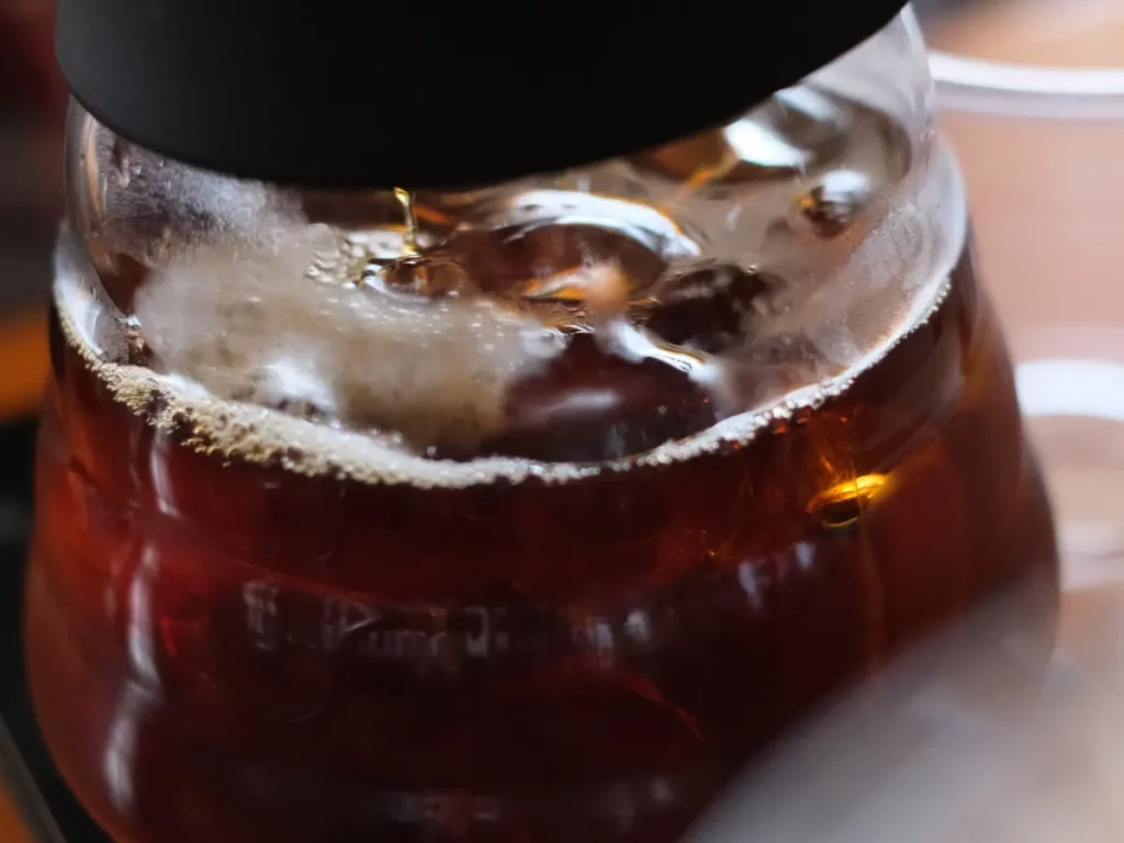 Close-up image of a glass container filled with freshly brewed coffee, showing bubbles and steam near the top. | Hustle Coffee