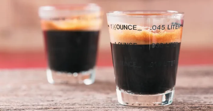 Two shot glasses filled with dark coffee are placed on a rustic wooden surface, with one glass in focus and the other slightly blurred. | Hustle Coffee