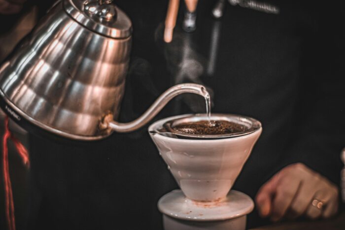 A person pours hot water from a metal kettle over coffee grounds in a ceramic dripper. | Hustle Coffee
