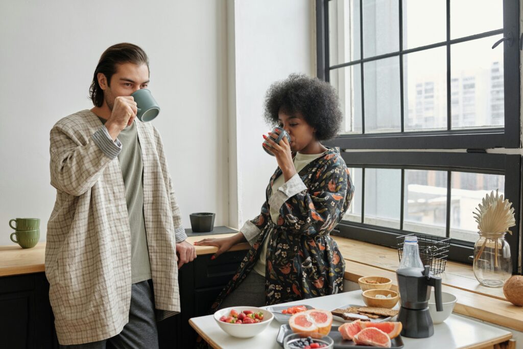 Two people drinking from mugs in a kitchen with breakfast food on the counter by a large window. | Hustle Coffee