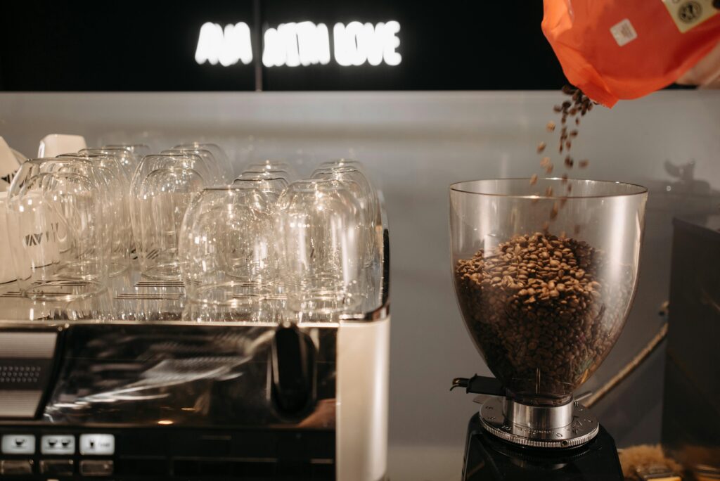 Coffee beans are being poured into a grinder next to stacked glass cups in a modern coffee shop setting. | Hustle Coffee