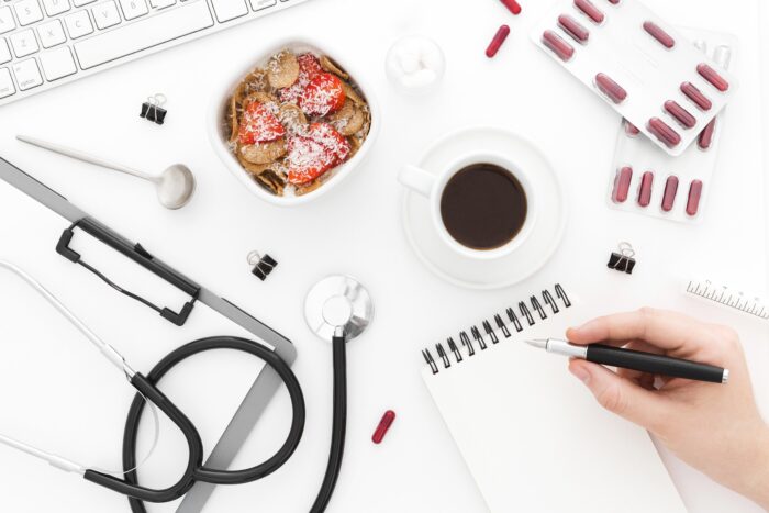 A hand writes in a notebook near coffee, cereal, pills, a stethoscope, and a computer keyboard on a white desk. | Hustle Coffee