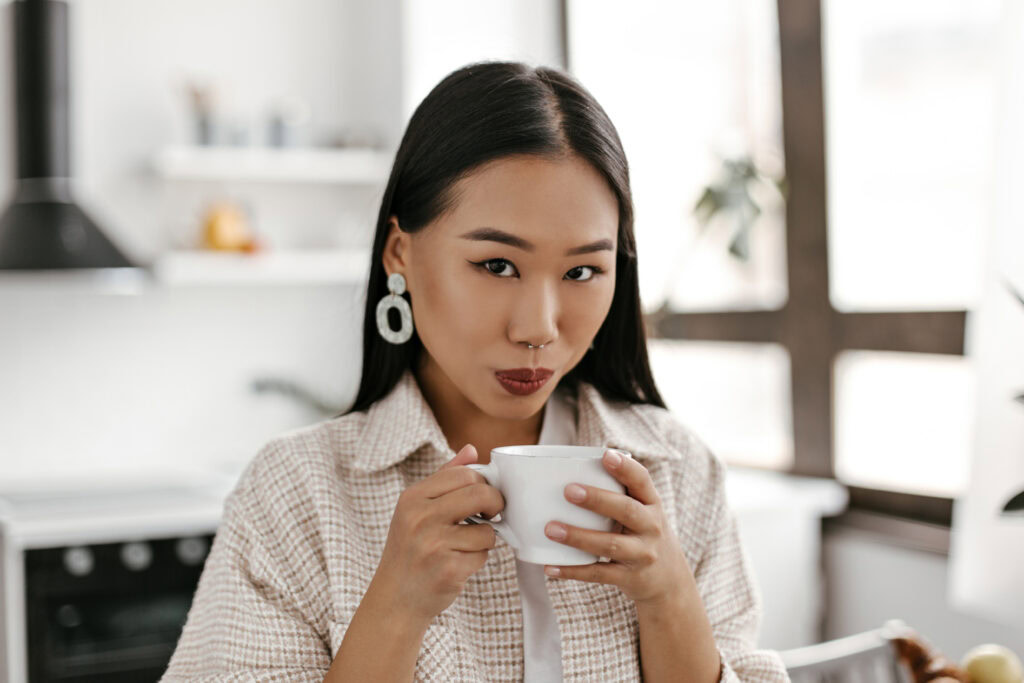 Woman with long dark hair sips coffe from a white mug in a bright kitchen, looking at the camera. | Hustle Coffee