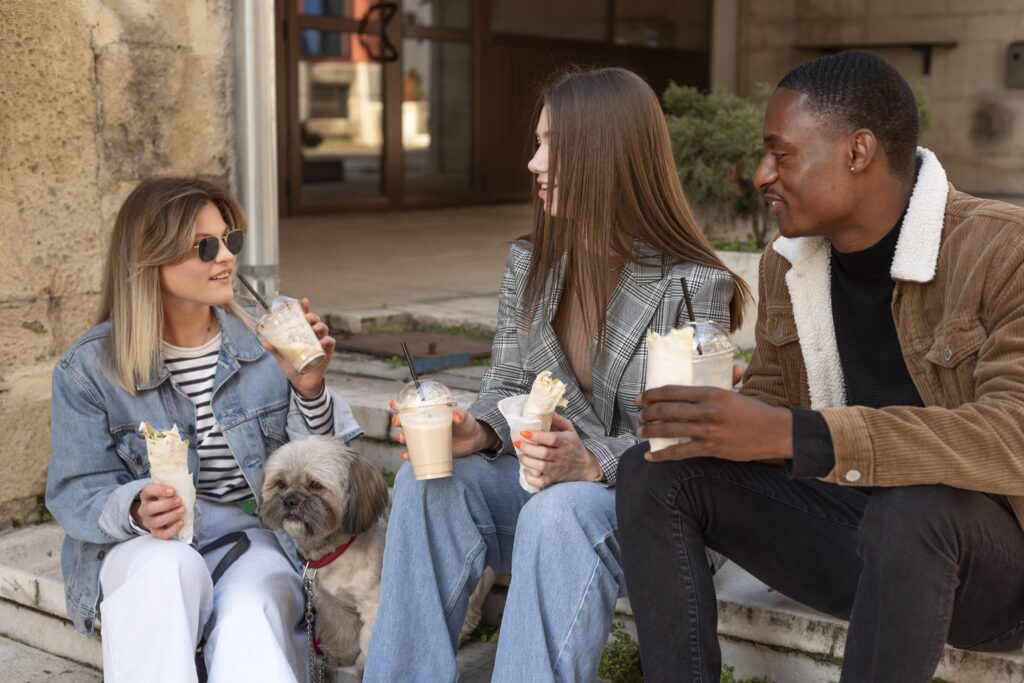 Three friends sit on outdoor steps with drinks, chatting and smiling, while a fluffy dog rests beside them. | Hustle Coffee