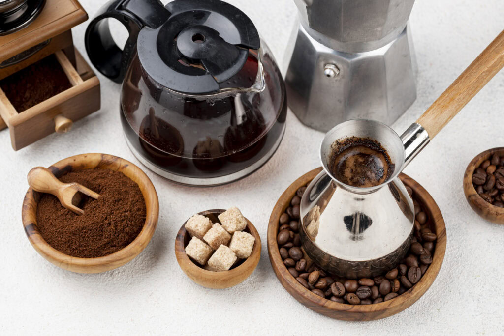 Coffee-making items including a cezve, coffee pot, moka pot, ground coffee, beans, and sugar cubes on a white surface. | Hustle Coffee