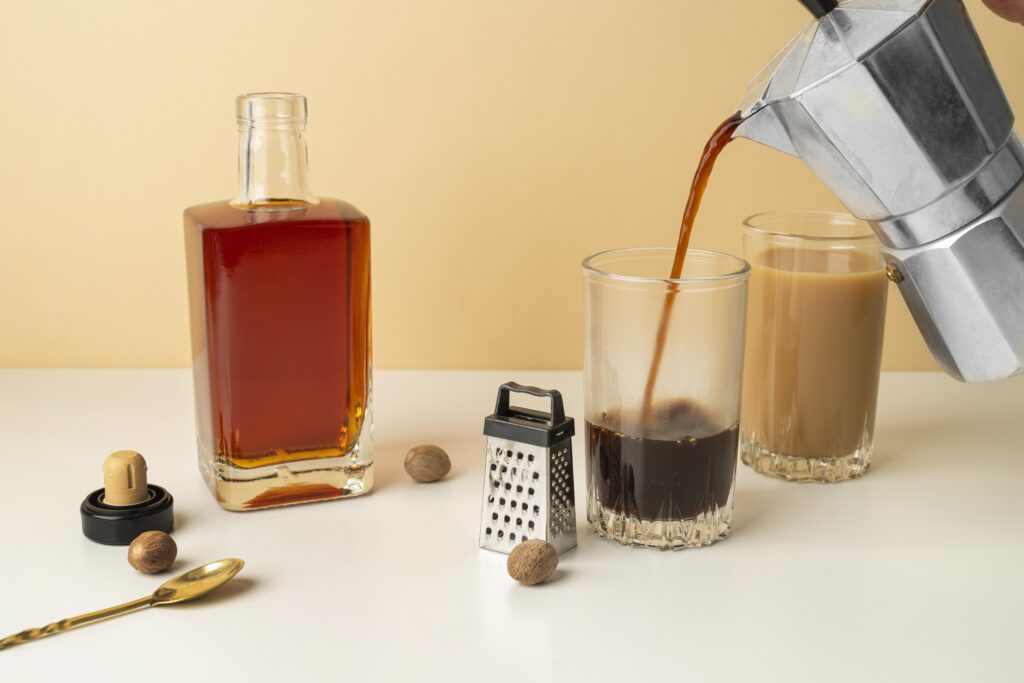 Coffee being poured into a glass, with a bottle, grater, nutmeg, and another coffee glass with milk on a white surface. | Hustle Coffee