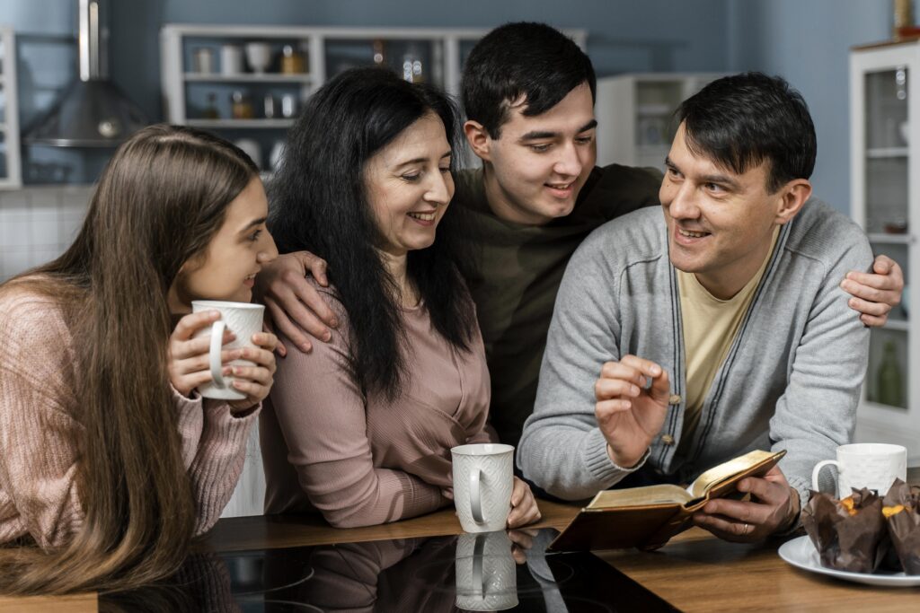 A family of four sits in a kitchen, smiling and talking, with mugs and an open book on the table. | Hustle Coffee