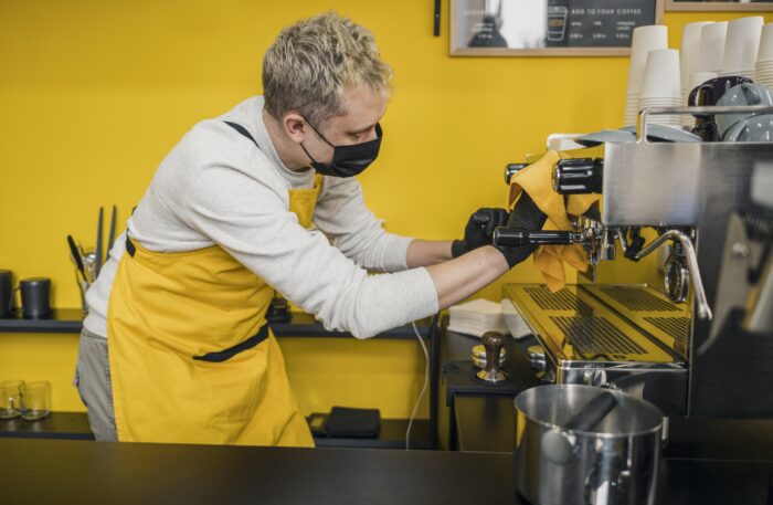 Barista wearing a yellow apron and mask cleans an espresso machine in a coffee shop with a bright yellow wall. | Hustle Coffee
