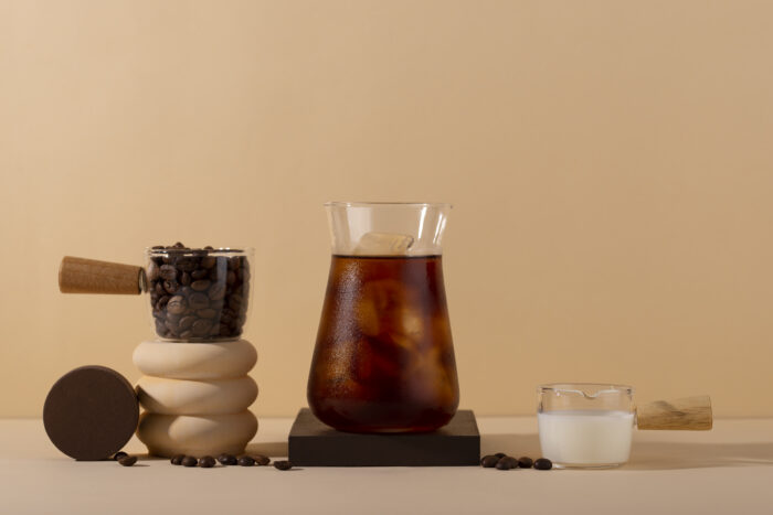 A glass of iced coffee, a cup of coffee beans, and a small glass of milk on a beige background. | Hustle Coffee