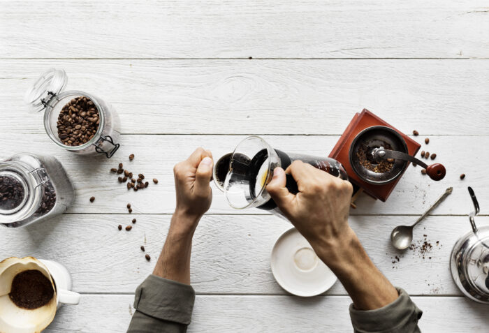 Person brewing coffee with a pour-over method, surrounded by coffee beans, grinder, and kettle on a white wooden table. | Hustle Coffee