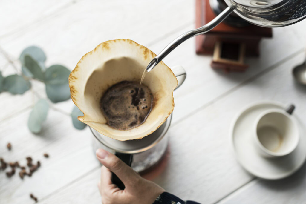 A hand pours hot water over coffee grounds in a filter, making pour-over coffee on a white wooden table. | Hustle Coffee