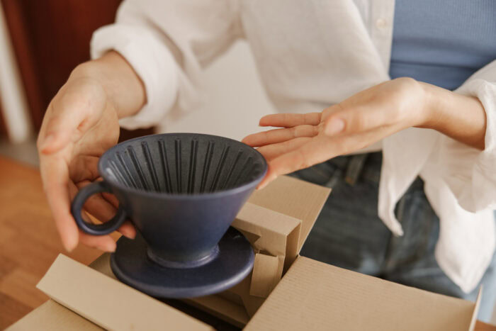 A person holds a dark blue ceramic coffee dripper above an open cardboard box on a table. | Hustle Coffee