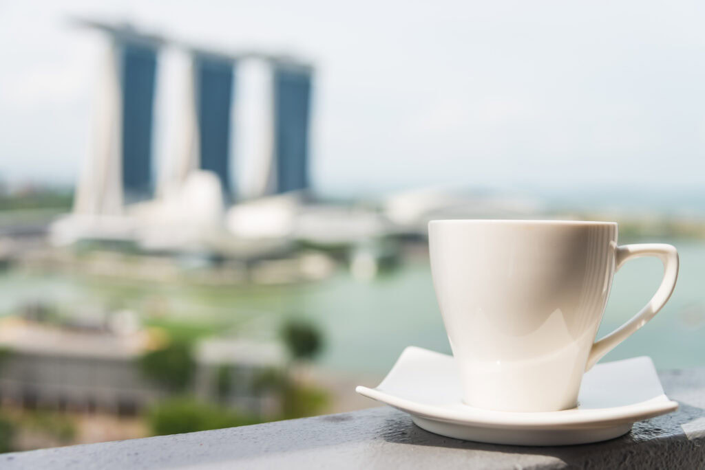 A white coffee cup on a saucer with a blurred view of Marina Bay Sands in Singapore in the background. | Hustle Coffee