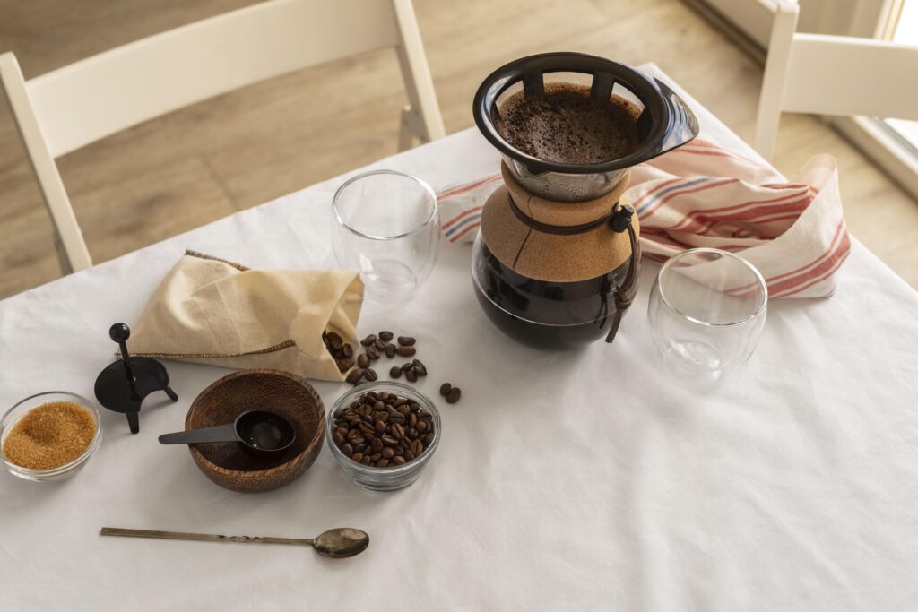 A coffee maker, two empty glasses, coffee beans, a spoon, and sugar sit on a white table with a striped cloth. | Hustle Coffee