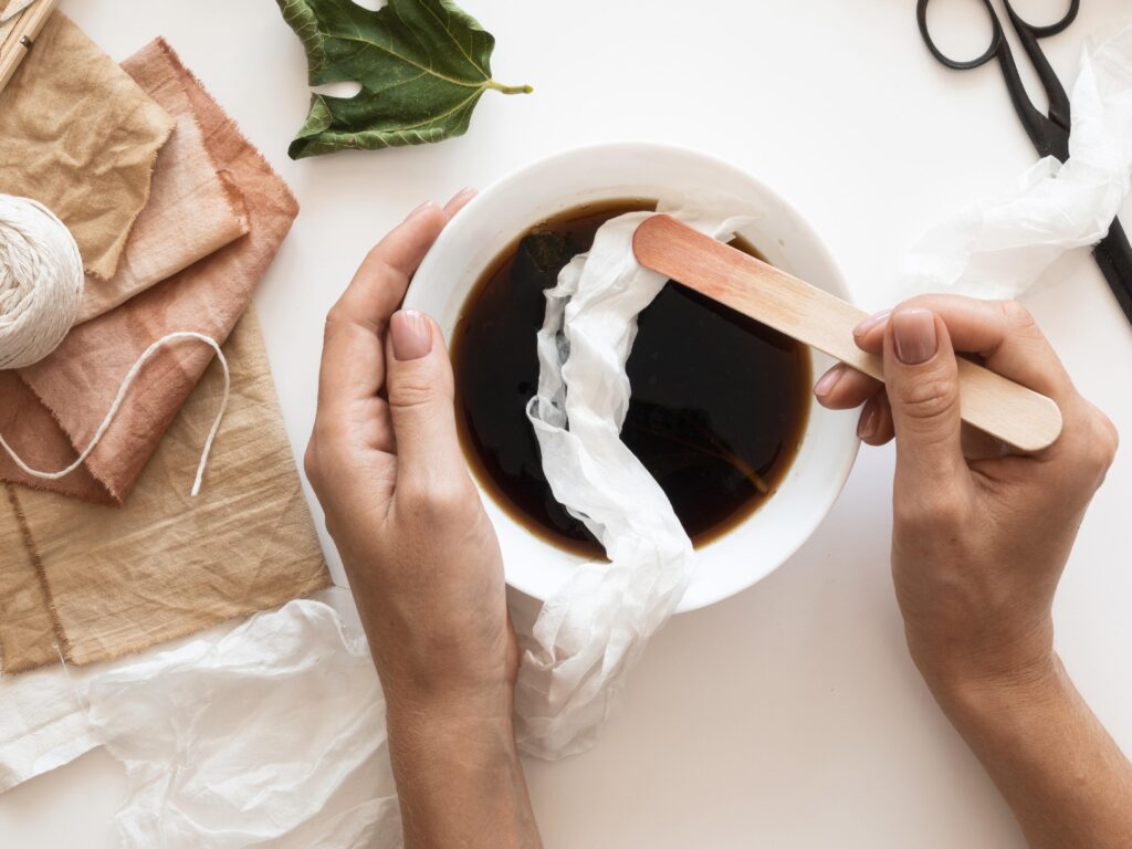 Hands dipping fabric into a bowl of dark liquid with craft supplies and a leaf nearby on a white surface. | Hustle Coffee