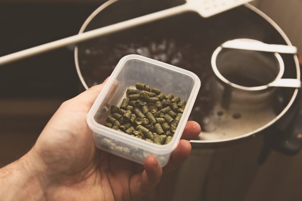 A hand holds a container of green hop pellets over a brewing pot with a slotted spoon and strainer inside. | Hustle Coffee
