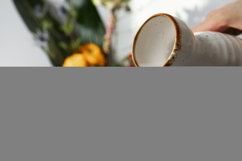 A hand holds a white ceramic mug with a brown rim, with blurred flowers and leaves in the background. | Hustle Coffee