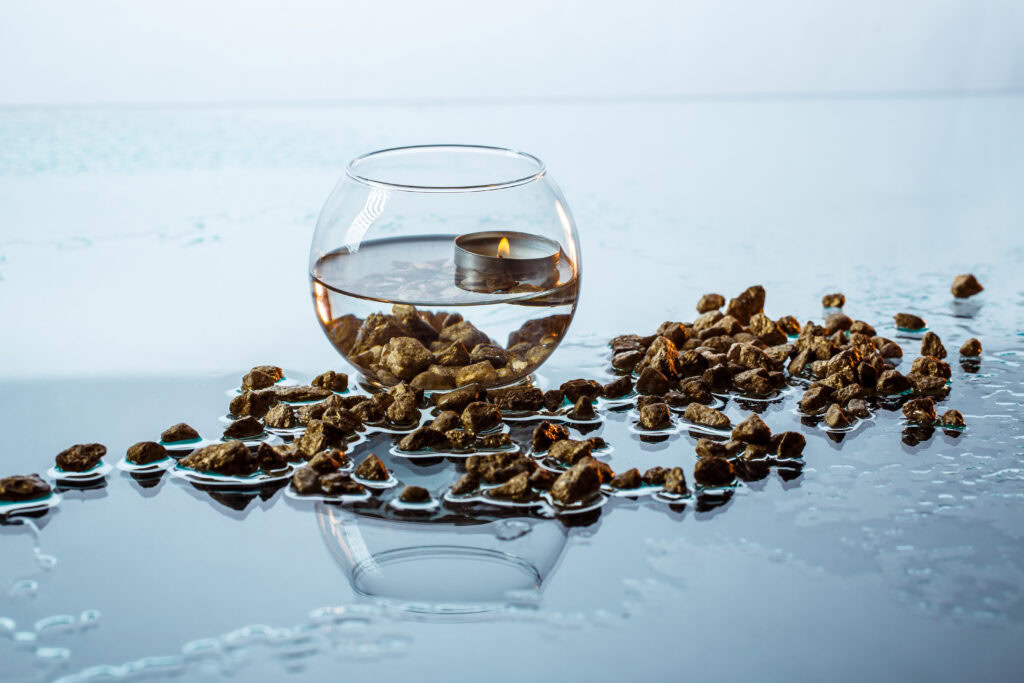 A floating candle in a glass bowl with water and stones, surrounded by scattered wet pebbles on a reflective surface. | Hustle Coffee