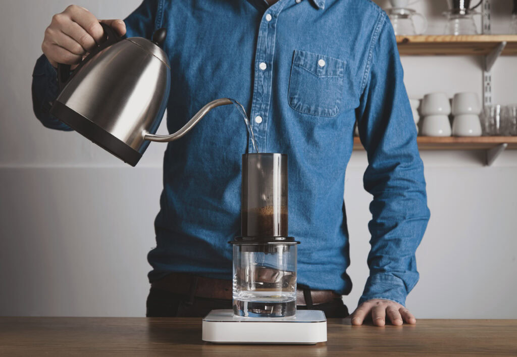 A person in a blue shirt pours hot water from a kettle into an Aeropress coffee maker over a glass on a scale. | Hustle Coffee