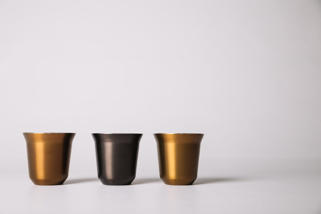 Three metallic coffee capsules—two gold, one black—are lined up on a white surface with a plain light background. | Hustle Coffee