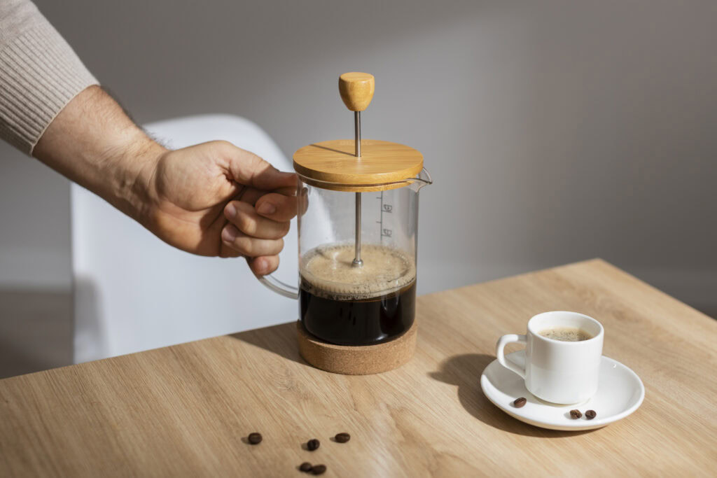 A person presses a French press filled with coffee next to a cup of espresso on a wooden table. | Hustle Coffee