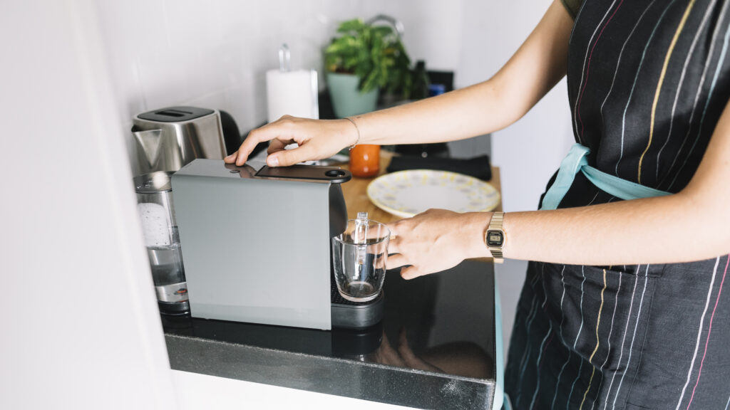 Person in apron making single-serve coffee with a pod machine, holding a cup under the dispenser on the kitchen counter. | Hustle Coffee