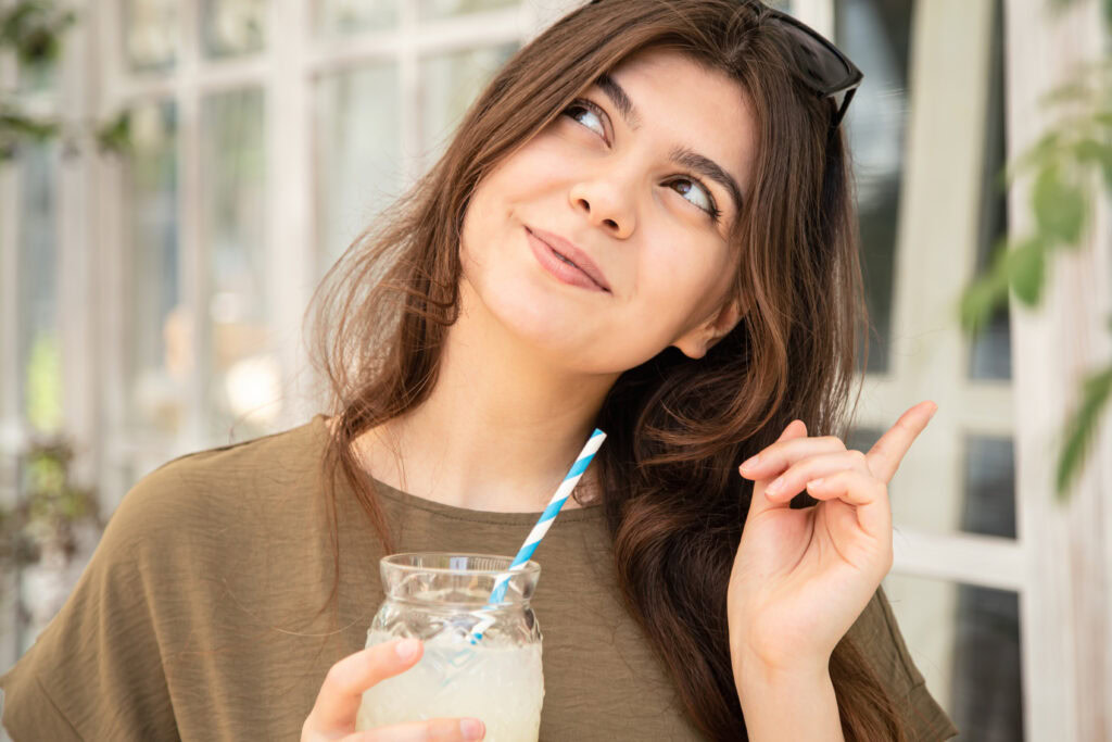 Young woman holding a drink and smiling, looking up thoughtfully, with her finger raised as if she has an idea. | Hustle Coffee