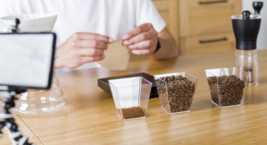 Person preparing coffee, with coffee beans, ground coffee, grinder, and brewing equipment on a wooden table. | Hustle Coffee