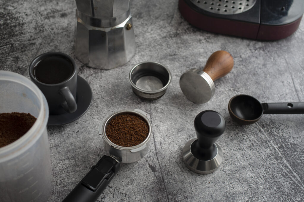 Various coffee-making tools and ground coffee on a gray surface, including a portafilter, tamper, and moka pot. | Hustle Coffee