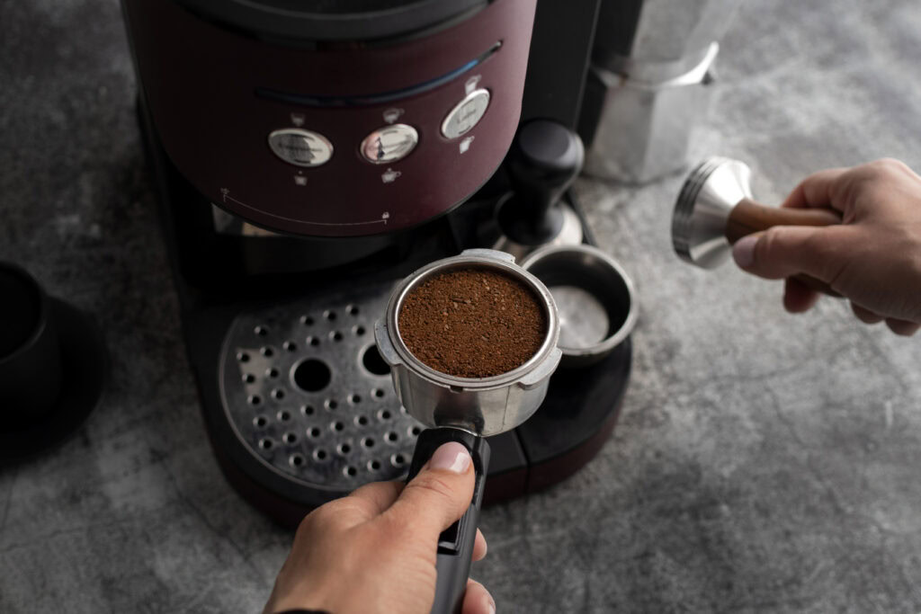 A person tamps ground coffee in a portafilter next to an espresso machine on a gray countertop. | Hustle Coffee