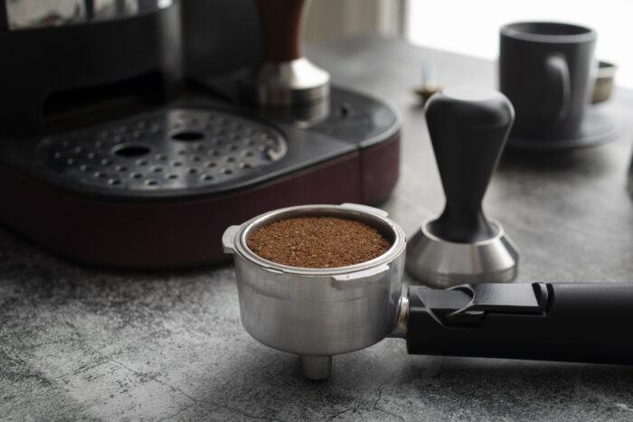 A portafilter filled with ground coffee sits on a countertop near an espresso machine and tamper. | Hustle Coffee