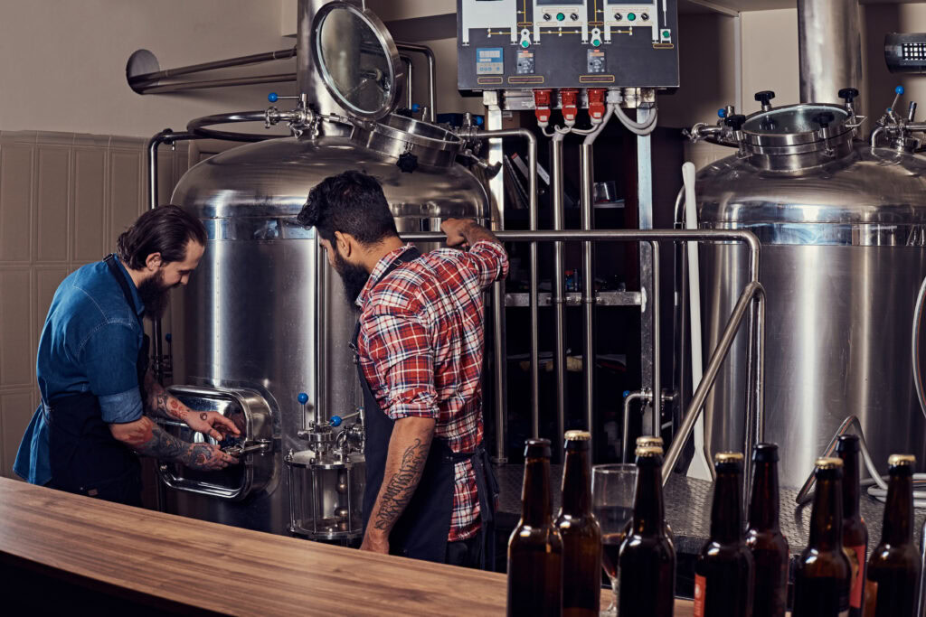 Two men in aprons inspect stainless steel brewing tanks in a brewery, with beer bottles on a counter in front. | Hustle Coffee