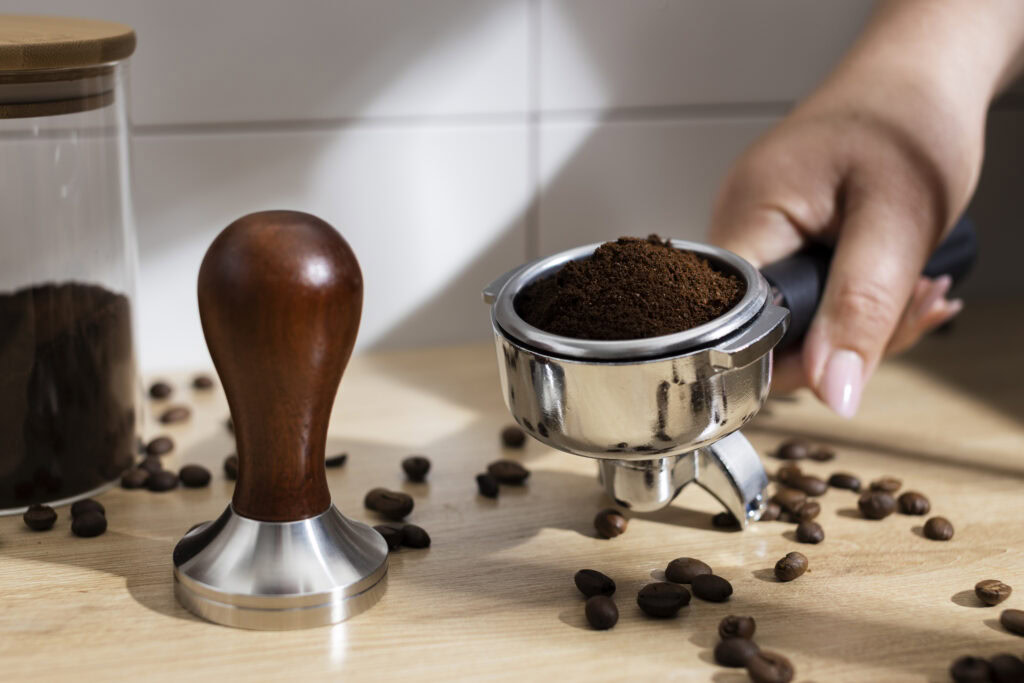A hand holds a portafilter filled with ground coffee next to a tamper, with coffee beans scattered on the table. | Hustle Coffee