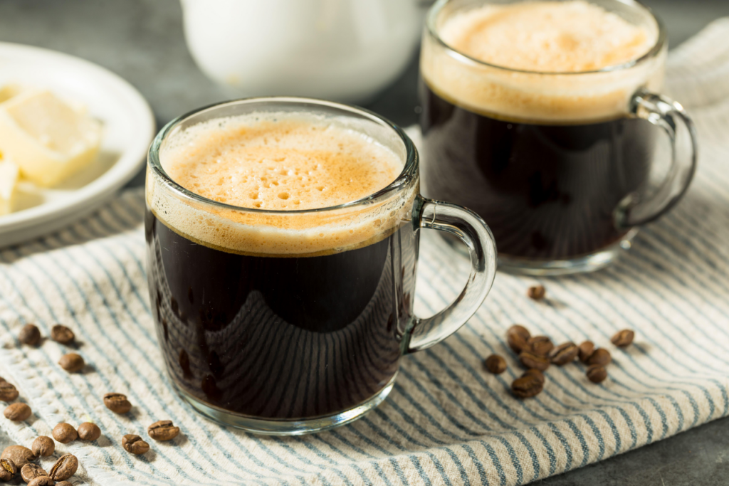 Two glass mugs of black coffee with frothy tops on a striped cloth, with coffee beans and a plate of butter nearby. | Hustle Coffee