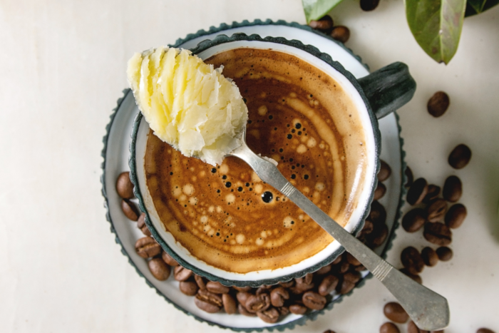 A cup of coffee with a spoonful of butter held above it, surrounded by coffee beans and green leaves. | Hustle Coffee