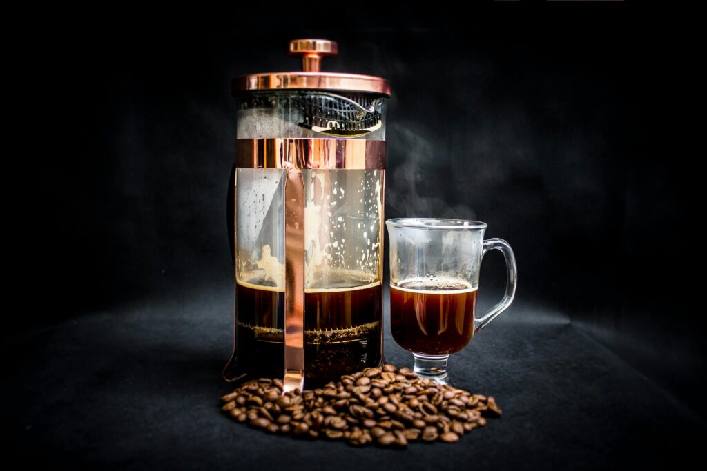 A French press with coffee, a glass mug of coffee, and coffee beans scattered on a dark surface. | Hustle Coffee