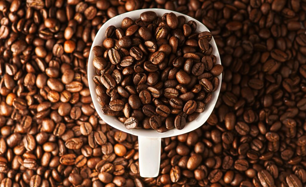 A white coffee cup filled with roasted coffee beans, surrounded by more coffee beans. | Hustle Coffee
