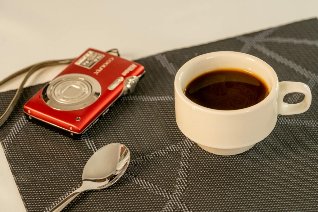 A white cup of black coffee, a spoon, and a red digital camera on a textured placemat. | Hustle Coffee