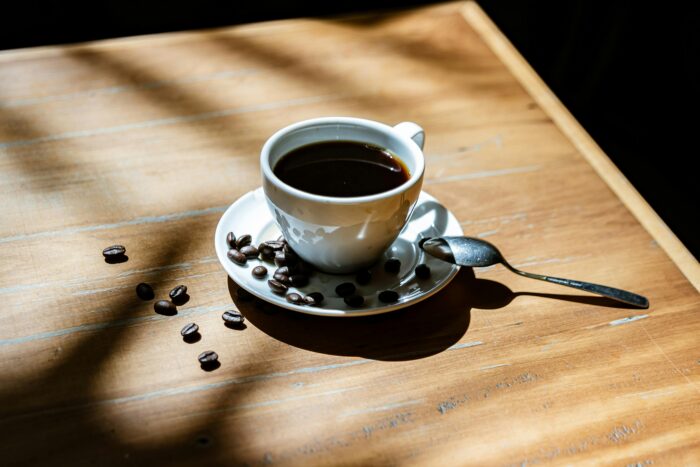 A cup of black coffee on a saucer with coffee beans and a spoon on a sunlit wooden table. | Hustle Coffee