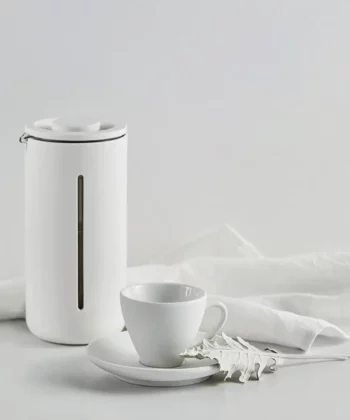 Minimalist white teapot next to a white teacup, saucer, and leaf-shaped spoon on a white cloth background. | Hustle Coffee