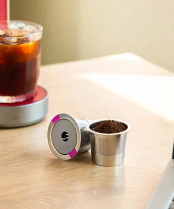 Reusable coffee pods filled with grounds sit on a wooden table next to a glass of iced coffee and a laptop. | Hustle Coffee
