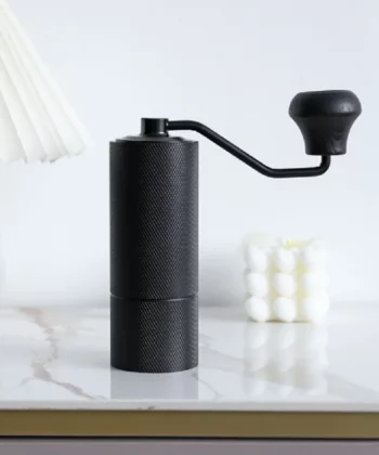 A black manual coffee grinder with a handle on a marble table near a white lamp and a decorative cube candle. | Hustle Coffee