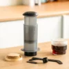 A coffee brewing device, scoop, stirrer, and a cup of coffee sit on a wooden counter in a kitchen setting. | Hustle Coffee
