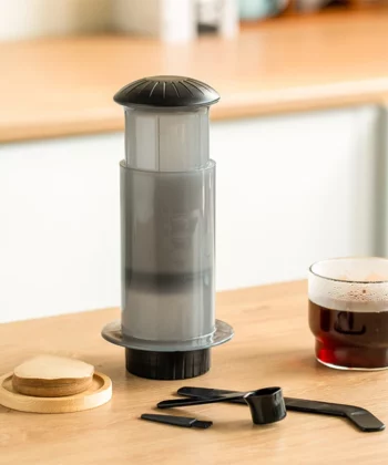 A coffee brewing device, scoop, stirrer, and a cup of coffee sit on a wooden counter in a kitchen setting. | Hustle Coffee