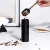 A hand pours coffee beans from a scoop into a black manual coffee grinder on a white table with books and decor. | Hustle Coffee