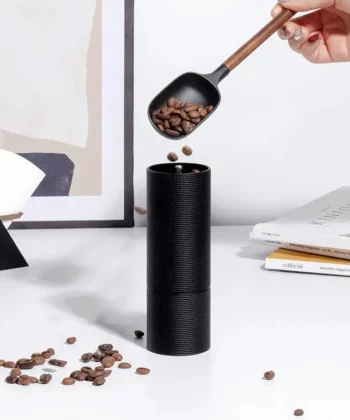A hand pours coffee beans from a scoop into a black manual coffee grinder on a white table with books and decor. | Hustle Coffee